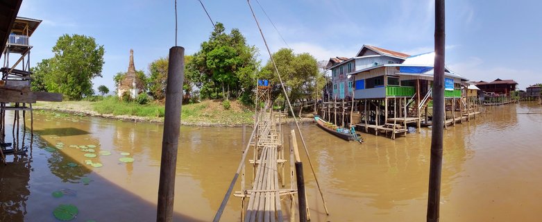 Bamboo bridge leading to colorful stilt houses over brown lake water with lotus pads and a pagoda visible in the background at Inle Lake, Myanmar