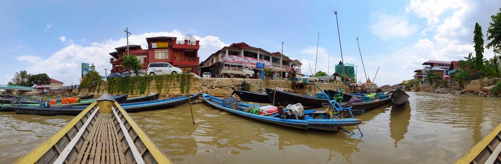 Multiple traditional wooden boats with blue painted hulls moored in brown water at Inle Lake, Myanmar, with multi-story buildings and vehicles visible on the shore under a blue sky with white clouds