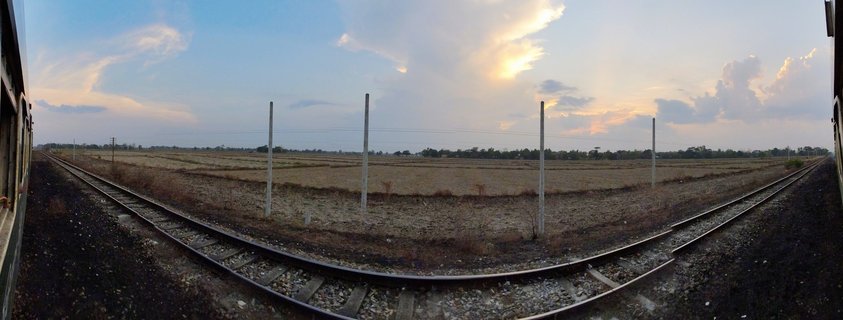 View from a moving train showing curved railway tracks through dry harvested fields in rural Myanmar at sunset with dramatic clouds and utility poles