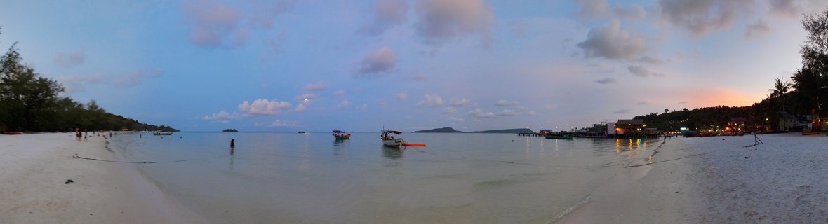 Panoramic beach scene at dusk showing white sand, calm shallow water with several traditional boats, tropical trees on both sides, illuminated buildings on the right, distant islands on the horizon, and a blue hour sky with scattered clouds and sunset colors