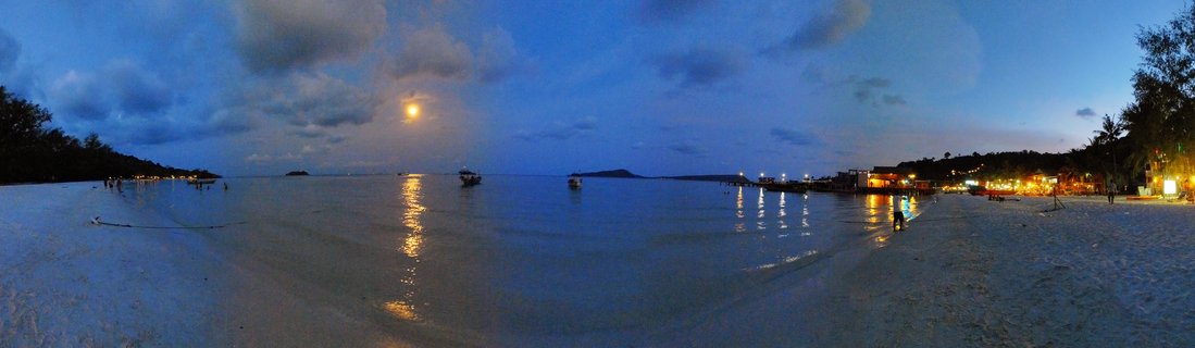 Panoramic beach scene at twilight showing calm ocean water with moon reflection, several boats anchored offshore, illuminated buildings along the shoreline, white sand beach, and cloudy blue sky with visible moon