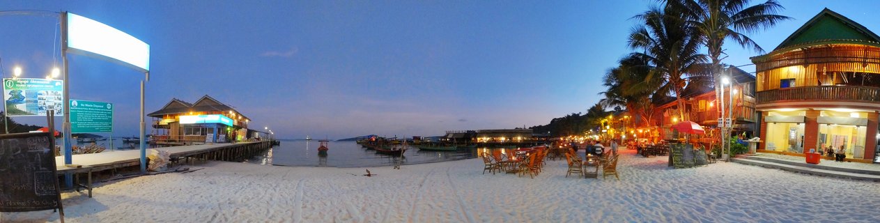 Panoramic beach scene at twilight showing wooden stilt buildings over water, palm trees, outdoor restaurant seating on white sand beach, anchored boats, and blue evening sky in Koh Rong, Cambodia