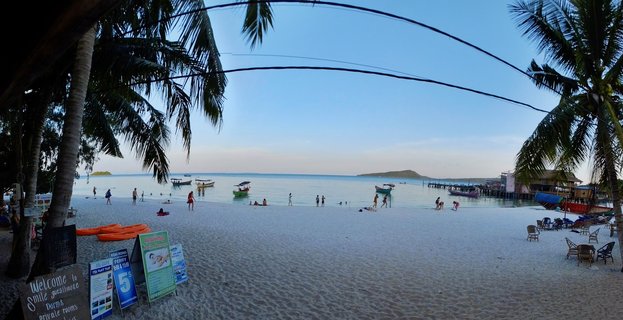 Wide beach scene showing white sand, palm trees, tourists swimming and walking, multiple boats in turquoise water, beach buildings on stilts, and promotional signs in foreground