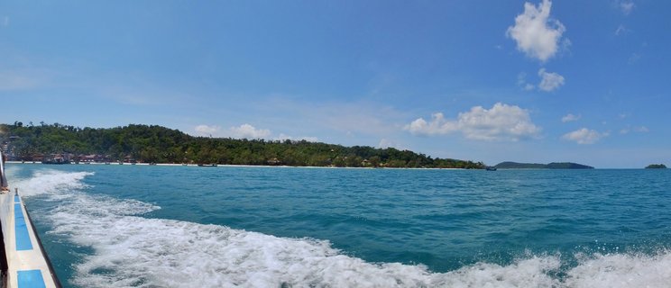 View from a moving ferry showing white wake in turquoise ocean water, with tropical Koh Rong island coastline featuring beaches, boats, and green hills in the background under blue sky