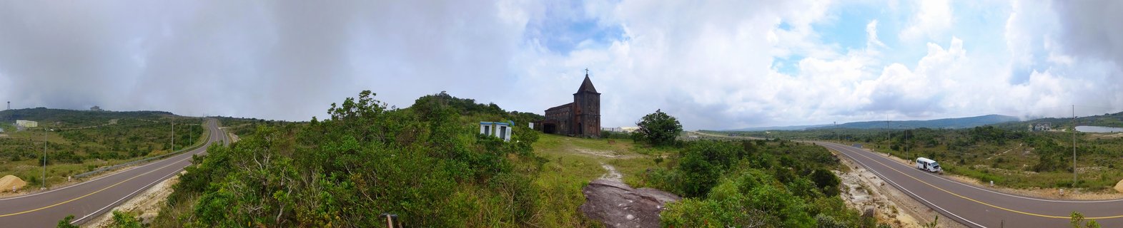 Panoramic view of a dark wooden church with steeple on a green hilltop between two curved roads under cloudy sky in rural Cambodia
