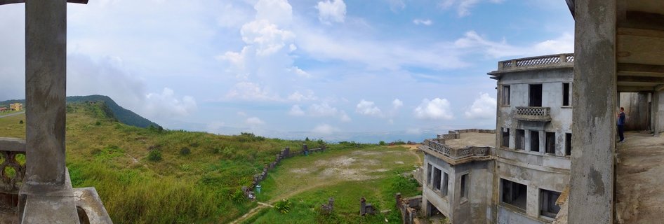 Abandoned multi-story concrete buildings on a green grassy hillside under blue sky with white clouds in Kampot, Cambodia