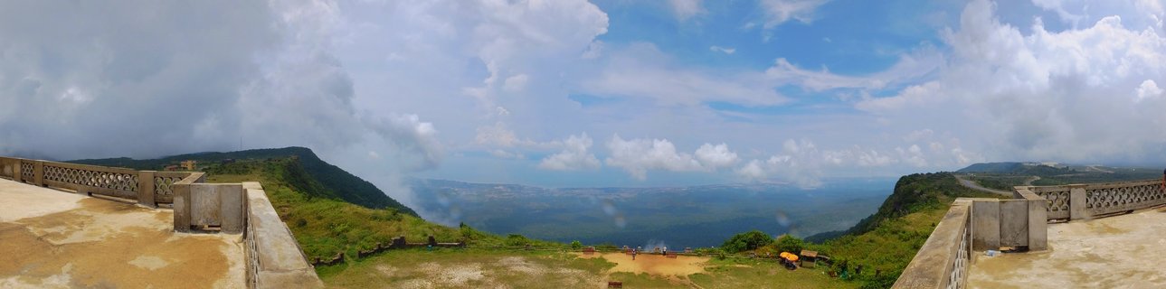 Panoramic landscape photograph from elevated concrete viewing platform with decorative railings on both sides, overlooking lush green tropical forest-covered mountains and expansive valley plains under dramatic blue sky with scattered white cumulus clouds, Bokor Mountain, Kampot Province, Cambodia