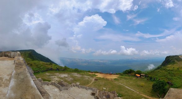 Panoramic landscape from Bokor Mountain viewpoint in Cambodia showing weathered French colonial concrete barrier ruins in the foreground, dense tropical jungle canopy covering rolling mountain slopes, distant valley plains through atmospheric haze, blue sky with scattered white cumulus clouds above.