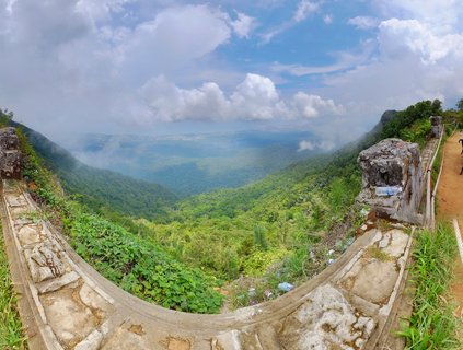 Panoramic view from a mountain viewpoint showing a curved stone wall in the foreground with lush green tropical forest filling the valley below under a blue sky with white clouds
