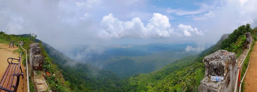 Panoramic mountain viewpoint showing a vast green forested valley with white clouds and mist, wooden benches and stone pillars in the foreground, Kampot Cambodia