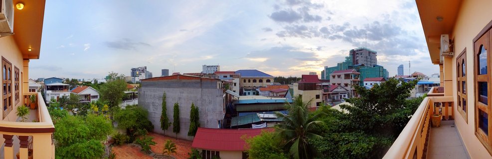Wide panoramic view of Phnom Penh city buildings and rooftops with tropical trees under cloudy evening sky, framed by cream-colored balcony railings