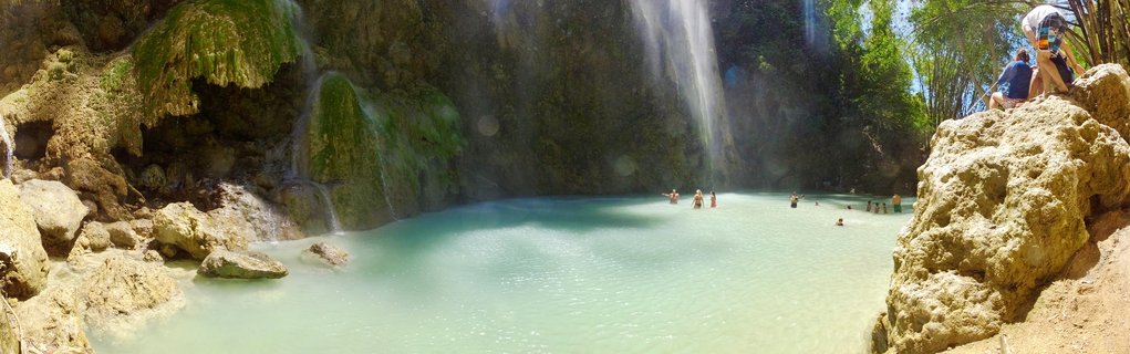 Wide view of a waterfall flowing into a turquoise natural swimming pool with people in the water and sitting on limestone rocks, surrounded by tropical vegetation