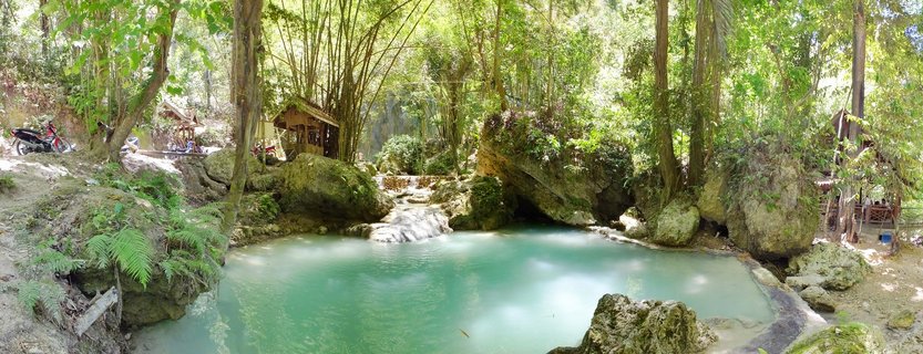 A natural turquoise spring pool surrounded by bamboo trees, large moss-covered rocks, and small wooden huts in a tropical forest