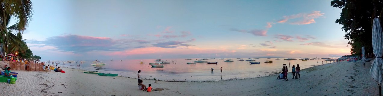 Panoramic beach scene at sunset in Bohol, Philippines showing white sand beach, calm ocean with multiple traditional outrigger boats, palm trees, and people walking and sitting along the shoreline under a pink and blue sky
