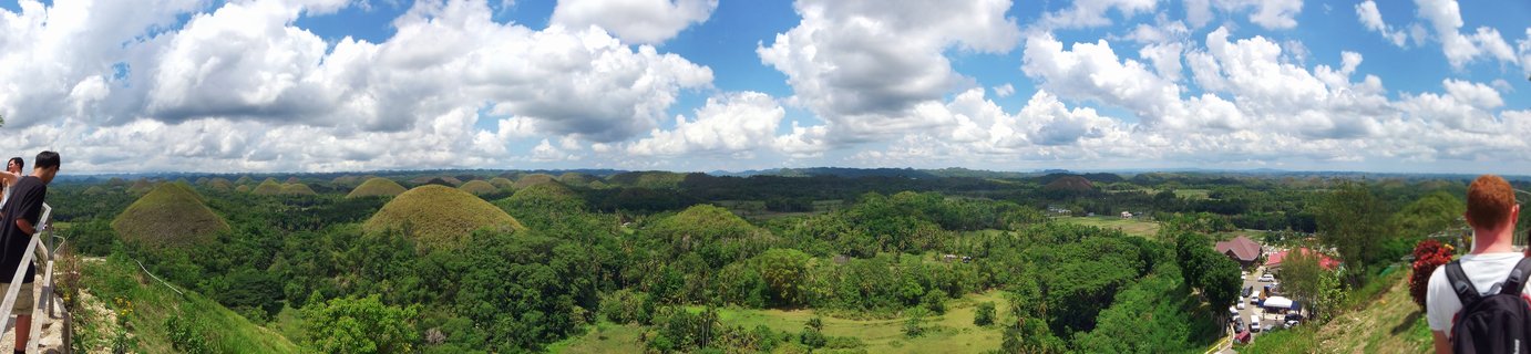 Panoramic view of numerous cone-shaped green hills across a valley landscape in Bohol, Philippines, with blue sky and white clouds above, tourists visible at viewing platform edges