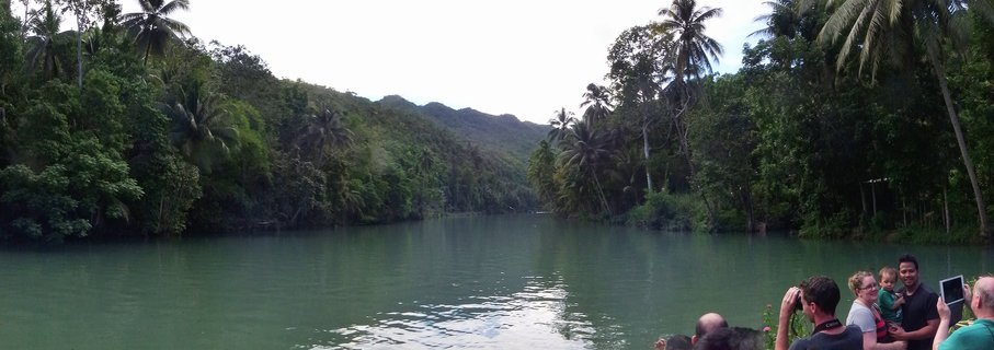 Wide panoramic view of Loboc River in Bohol with tourists on a boat in the foreground, tropical palm trees and dense green vegetation lining both riverbanks, and forested hills in the background under an overcast sky