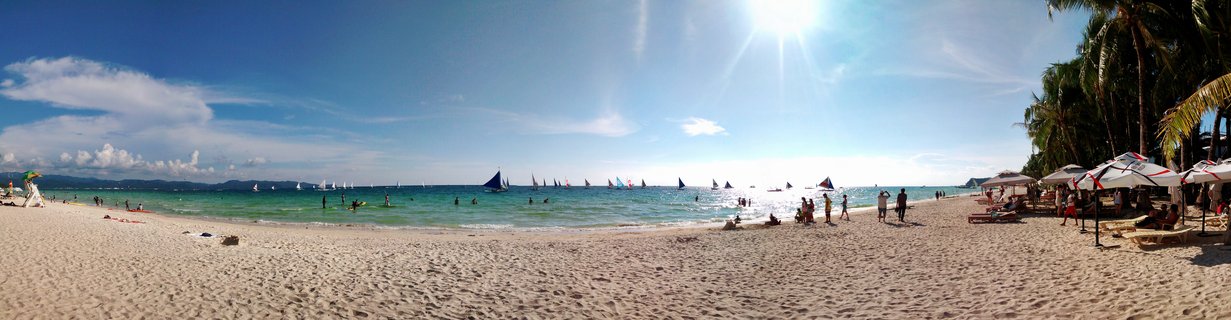 Panoramic view of White Beach Boracay showing white sand beach, turquoise ocean, multiple sailboats, beach umbrellas, palm trees, and tourists under bright sunny sky