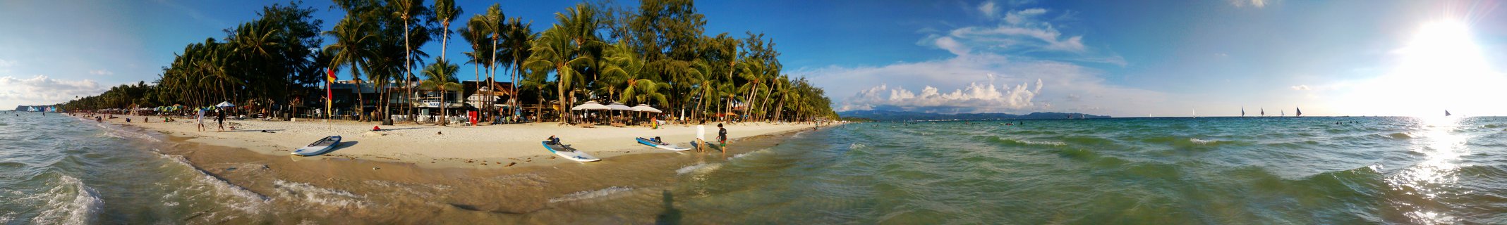 Wide panoramic photograph of a tropical beach showing white sand, turquoise ocean water, a row of tall palm trees along the shore, small boats on the beach, people walking, beach umbrellas, and sailboats visible in the distance under a blue sky with scattered clouds and bright sunlight.