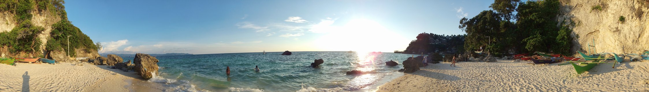 Panoramic beach scene showing white sand shore with traditional outrigger boats, people swimming in turquoise water, rocky cliffs with vegetation on both sides, and bright sun low on horizon over ocean