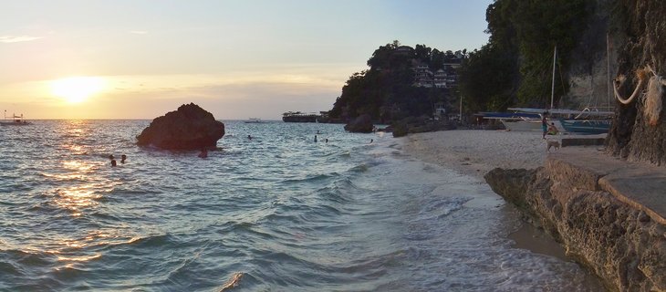 Beach sunset scene showing people swimming in turquoise ocean water with large rock formation, traditional outrigger boats docked on white sand beach, and resort buildings on vegetated cliffside