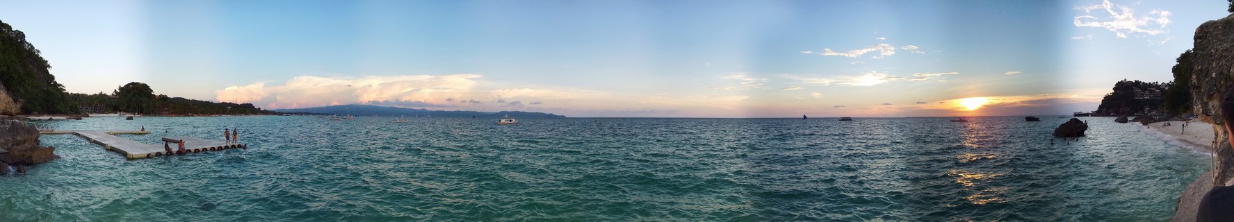 Panoramic seascape showing turquoise ocean water at sunset with a floating dock, boats, rocky coastline, and distant mountains in Boracay, Philippines