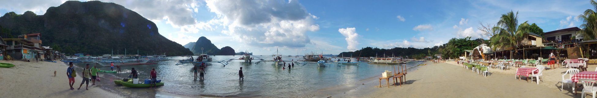 Panoramic view of a tropical beach in El Nido, Palawan with traditional Filipino outrigger boats in shallow water, limestone mountain formations in the background, beach resorts with tables and chairs, and people walking along the shore