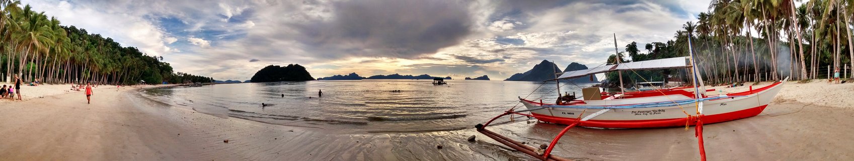 Panoramic view of tropical beach in Palawan Philippines with traditional red and white outrigger boat on white sand, palm trees, people swimming, and limestone islands in background under cloudy sky