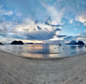 Wide beach scene showing white sand in foreground, calm reflective water, multiple limestone karst islands in the distance under a cloudy sunset sky with golden and blue tones