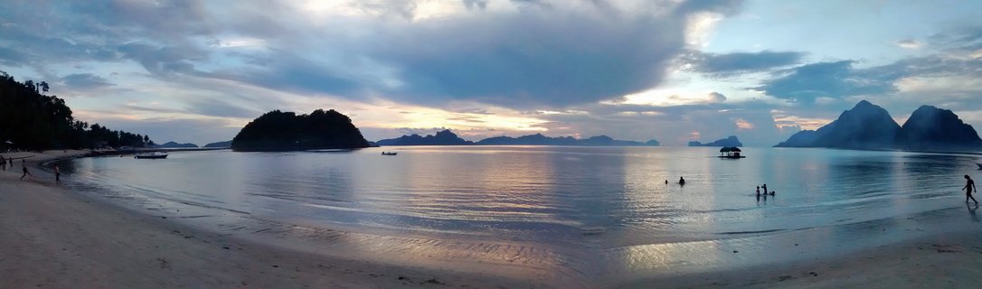Panoramic sunset view of tropical beach bay with limestone islands, calm water reflections, and people on shoreline in Palawan, Philippines