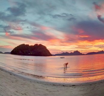Person bending over in shallow ocean water at sunset with forested limestone island and mountains in background, pink and orange clouds in sky, white sand beach in foreground