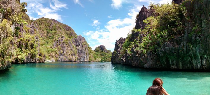 Tropical lagoon with turquoise water surrounded by steep limestone cliffs covered in green vegetation under blue sky with white clouds, two people visible in foreground