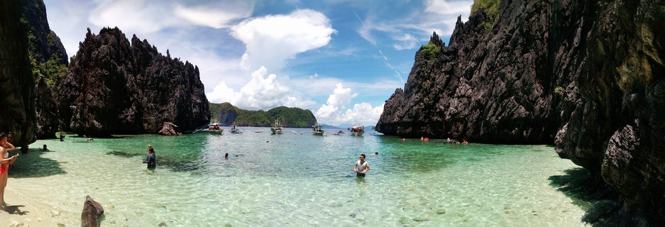 Panoramic view of tourists swimming in clear turquoise water between tall dark limestone rock formations in a tropical lagoon in the Philippines