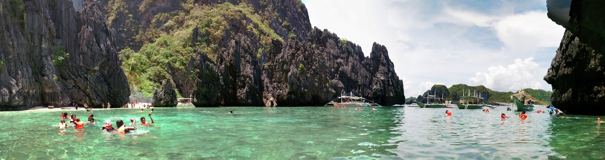 Aerial panoramic view of tourists swimming in clear turquoise tropical water with limestone cliffs, traditional boats, and white sand beach in El Nido, Palawan, Philippines