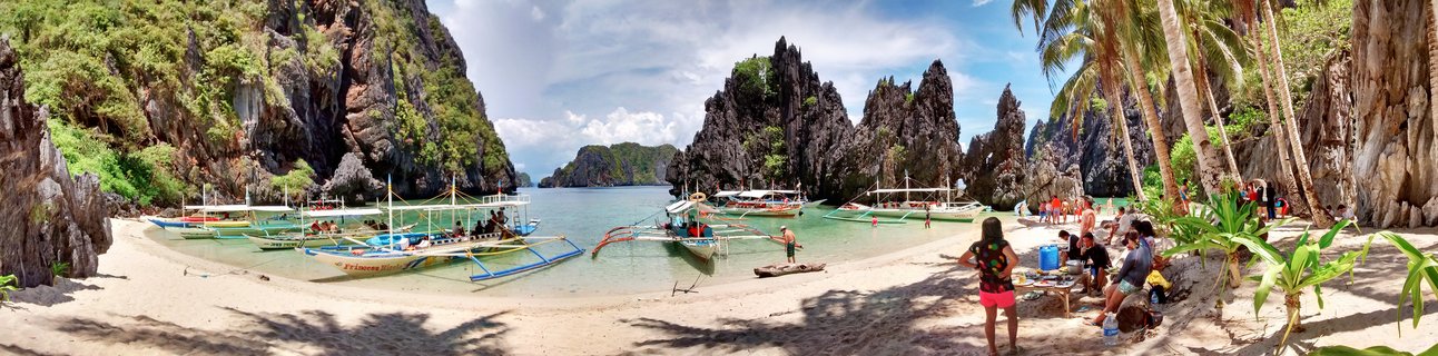 Wide panoramic view of a tropical beach bay in Palawan Philippines with limestone karst cliffs, traditional outrigger boats in turquoise water, palm trees, and tourists on white sand beach