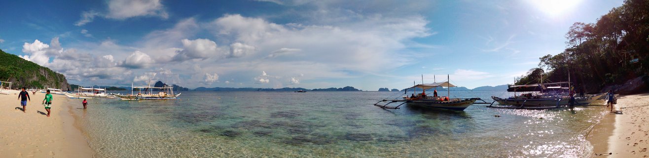 Panoramic view of a tropical beach in Palawan with traditional Filipino outrigger boats in clear turquoise water, white sand beach with people walking, limestone cliffs and blue sky with clouds