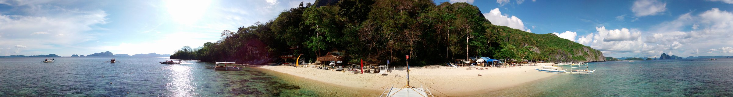 Panoramic tropical beach scene showing white sand, clear turquoise water, traditional outrigger boats, green limestone cliffs, and blue sky with clouds in Palawan, Philippines