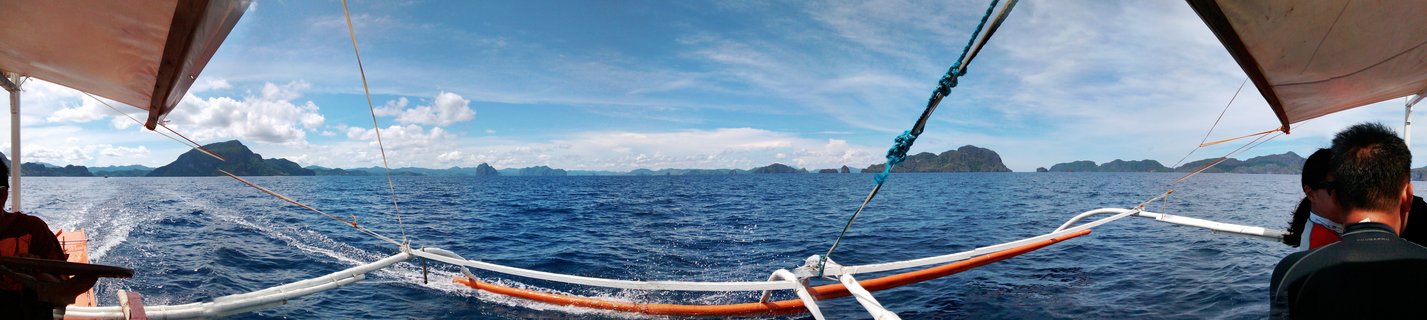 View from a traditional Filipino outrigger boat showing white and orange bamboo stabilizers, blue ocean water with boat wake, distant limestone islands, and passengers seated under canvas canopy