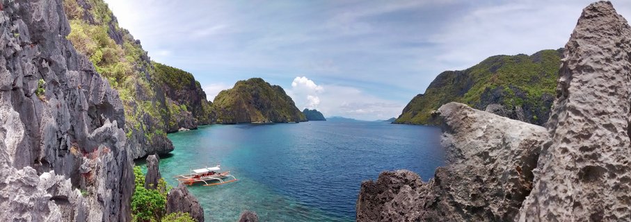 Panoramic view of a tropical bay in Palawan with limestone cliffs on both sides, turquoise water, and a traditional Filipino outrigger boat anchored in the foreground