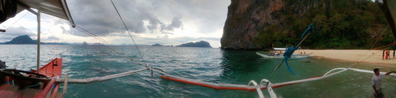View from a traditional Filipino outrigger boat showing turquoise tropical water, white sand beach, limestone cliffs covered in vegetation, and other bangka boats near shore in Palawan, Philippines