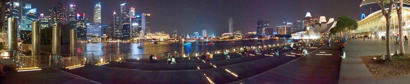 Night panoramic view of Singapore's Marina Bay showing illuminated skyscrapers across the water with people on waterfront promenade and the ArtScience Museum visible