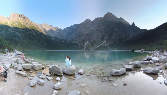 Panoramic view of Morskie Oko alpine lake with turquoise water, surrounded by rocky Tatra Mountain peaks, with visitors sitting on boulders along the shoreline under clear blue sky