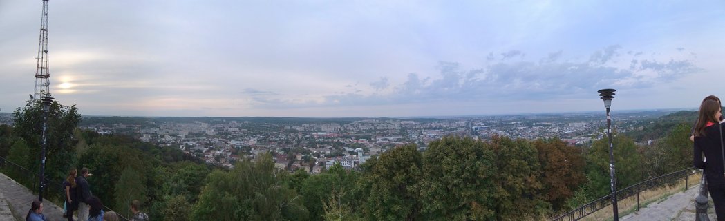 Wide panoramic photograph showing Lviv city skyline viewed from an elevated hilltop position with trees in foreground, people at viewing areas, and urban buildings stretching to horizon under cloudy sky