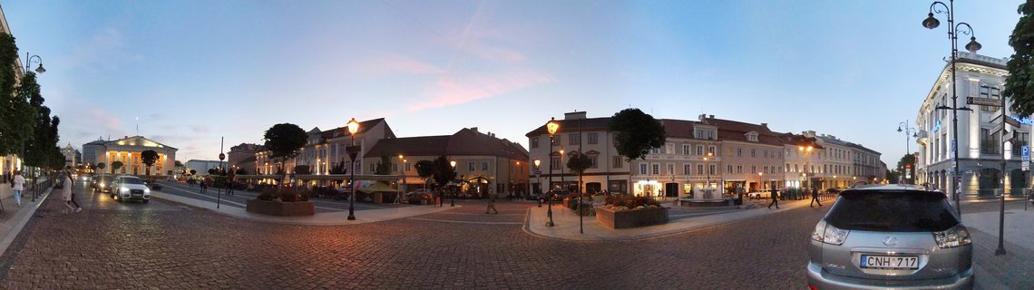 Panoramic street view of Vilnius Old Town square at dusk with illuminated historic buildings, cobblestone streets, street lamps, parked cars, and pedestrians walking
