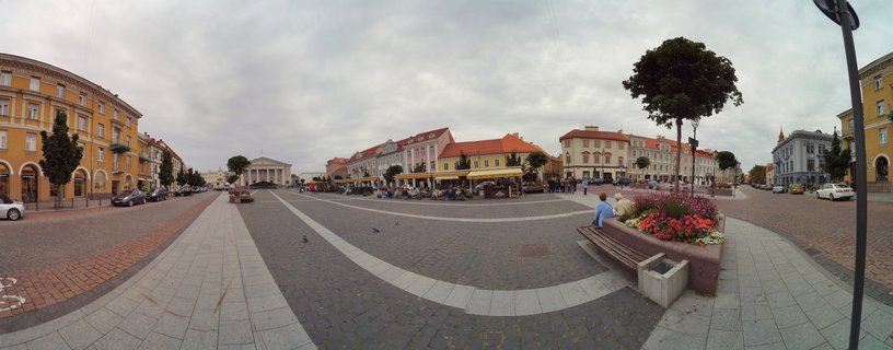 Wide panoramic view of Cathedral Square in Vilnius showing the white neoclassical cathedral with columns, colorful historic buildings with orange roofs, cobblestone plaza, outdoor cafés, trees, flower beds, and people walking