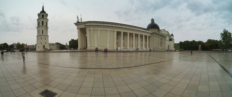 Wide view of Vilnius Cathedral with its colonnade and Bell Tower on Cathedral Square, with people walking across the paved plaza under cloudy skies