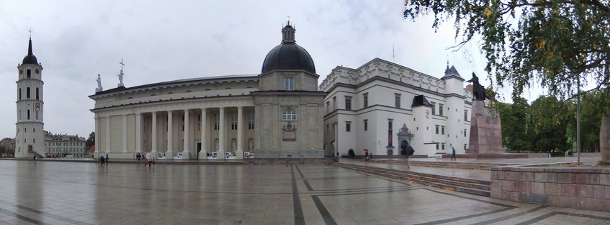 Wide view of Vilnius Cathedral Square showing the white Cathedral with columns and dome, tall Bell Tower on the left, Palace of Grand Dukes on the right, wet stone pavement, overcast sky, pedestrians with umbrellas