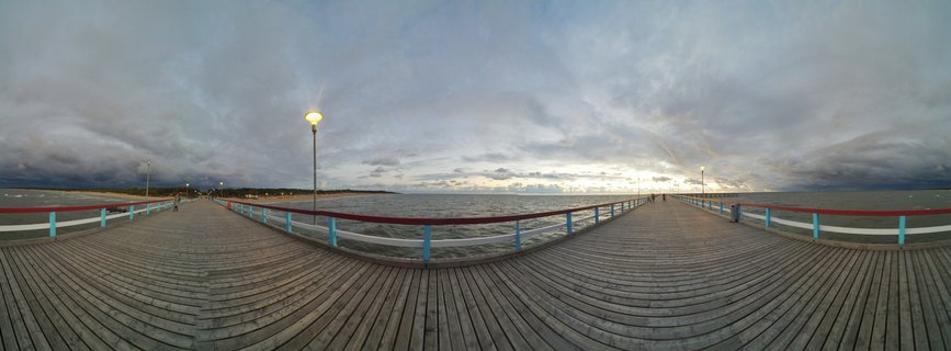 Wooden pier with blue and red railings extending over Baltic Sea waters under cloudy twilight sky with illuminated street lamps in Palanga, Lithuania