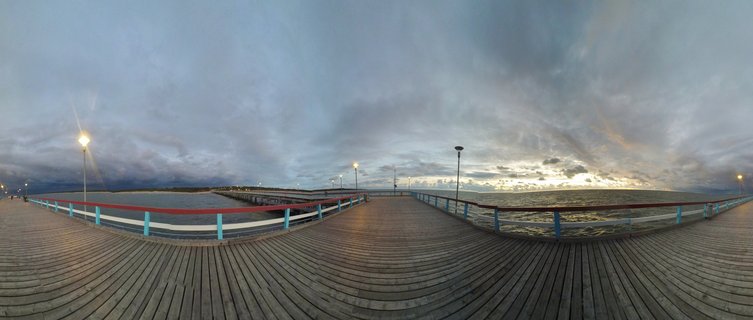 360-degree panoramic view of a wooden pier bridge extending over Baltic Sea waters in Palanga, Lithuania, with blue and white railings, illuminated street lamps, and cloudy evening sky