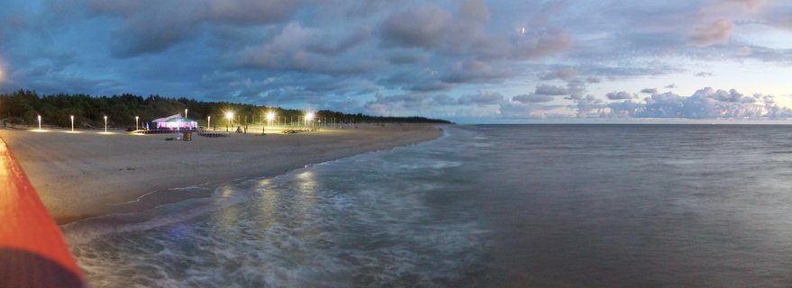 Panoramic view of Palanga beach in Lithuania at dusk showing illuminated beach area with lights, sandy shore, Baltic Sea waves, tree line, cloudy twilight sky with moon visible