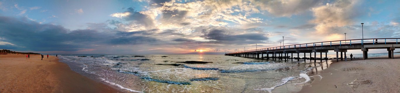Panoramic view of Palanga Pier extending into the Baltic Sea at sunset with golden light reflecting on water, waves on sandy beach, and people walking along shore and pier